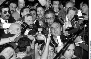 Black and white photo of crowd of press holding out microphones to a few people in the middle of them